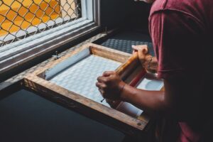 Artisan manipulant un cadre de sérigraphie près d’une fenêtre grillagée